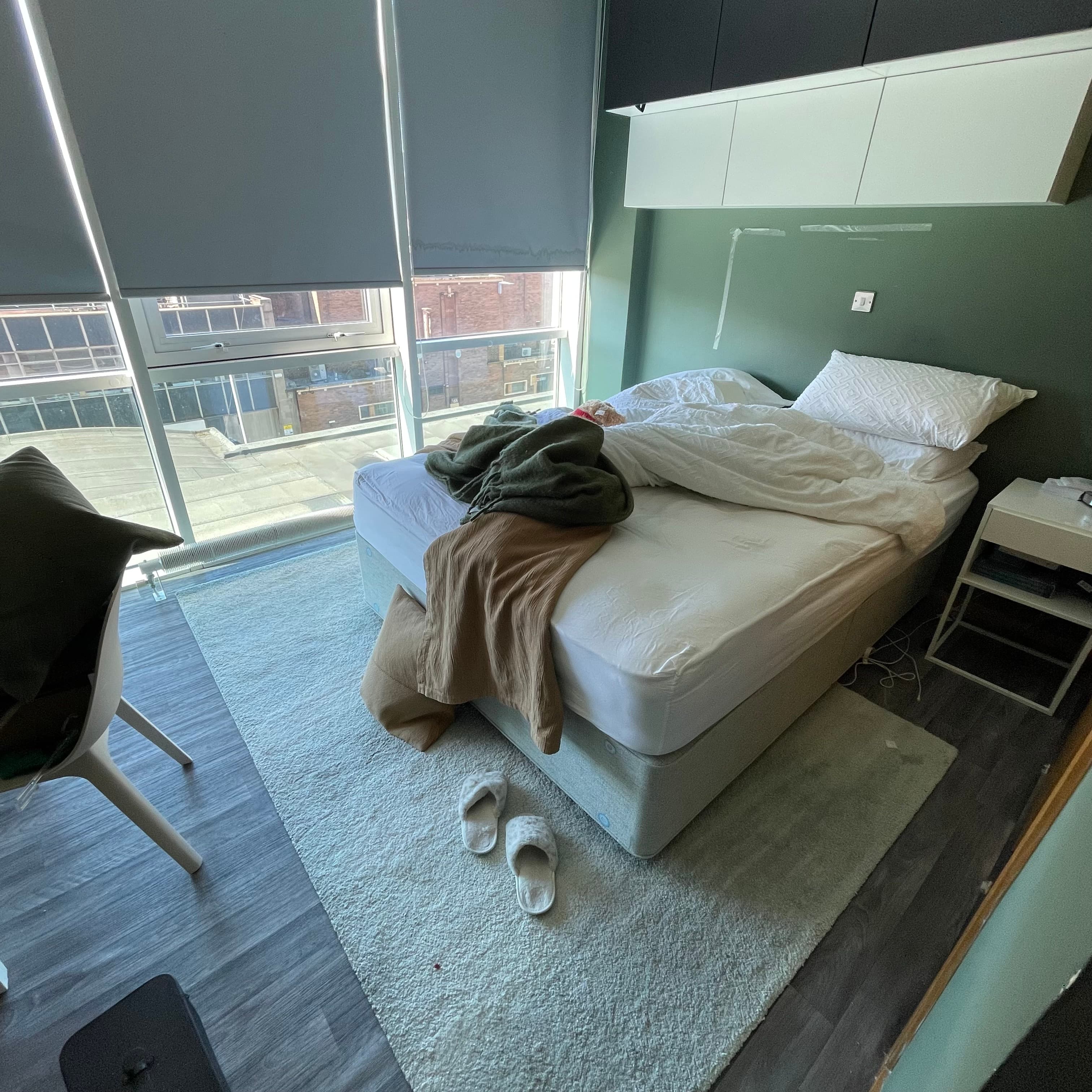 Unmade bed with white bedding and green blankets in a modern room with large windows.