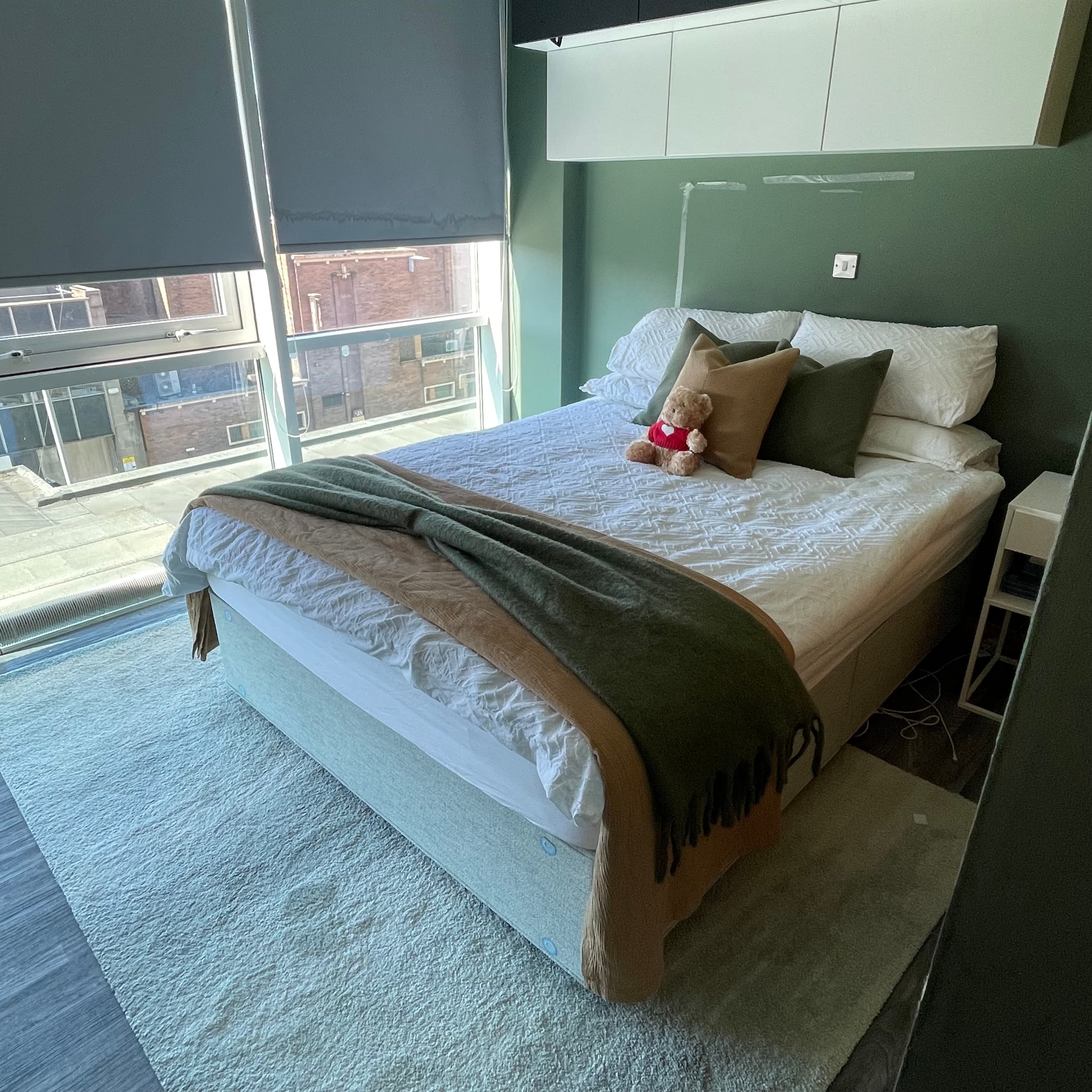Modern bedroom with white textured bedding, green pillows, a teddy bear, and a large window.